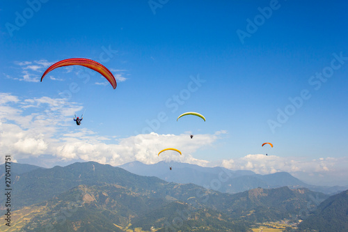 four paragliders on multi-colored parachutes fly over a green mountain valley against a blue sky with white clouds, aerial view