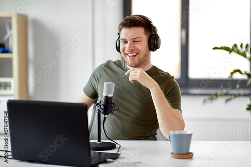 technology, mass media and podcast concept - happy young male audio blogger in headphones with laptop computer and microphone broadcasting at home office photo