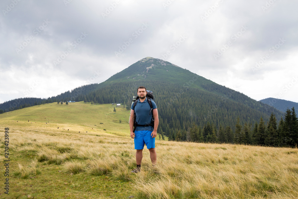 Young handsome man with backpack standing in mountain grassy valley on copy space background of summer woody mountain peak and blue sky. Active lifestyle, sports and recreation concept