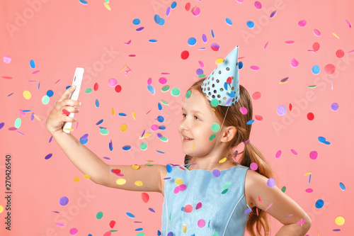 Cheerful little girl celebrates birthday. The child holds the phone, takes a selfie in the rain of confetti. Closeup portrait on pink background.