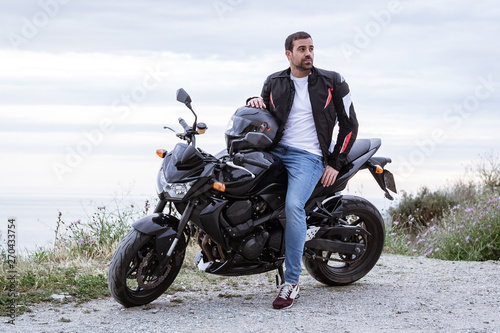 Young man biker with his black motorbike ready to drive, in front of the sea