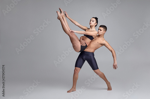 The couple of a young modern ballet dancers in black suits are posing over a gray studio background.