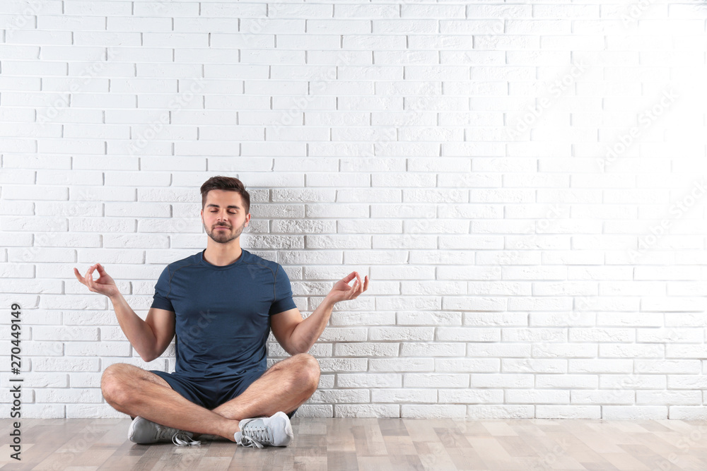 Handsome young man sitting on floor and practicing zen yoga near brick wall, space for text