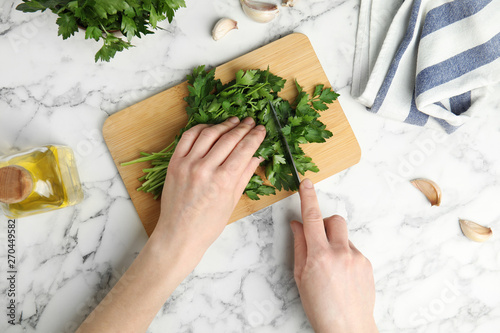 Woman cutting fresh green parsley on wooden board at marble table, top view photo