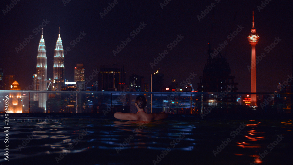 Night view of woman bathing in rooftop swimming pool and looking at the city with illuminated Petronas Towers and Kuala Lumpur Tower, Malaysia