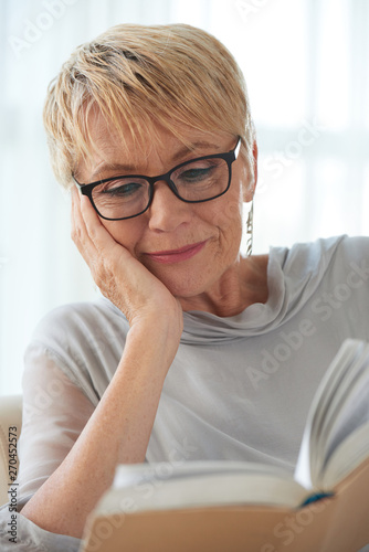 Mature beautiful woman wearing glasses keen on reading an interesting book at home