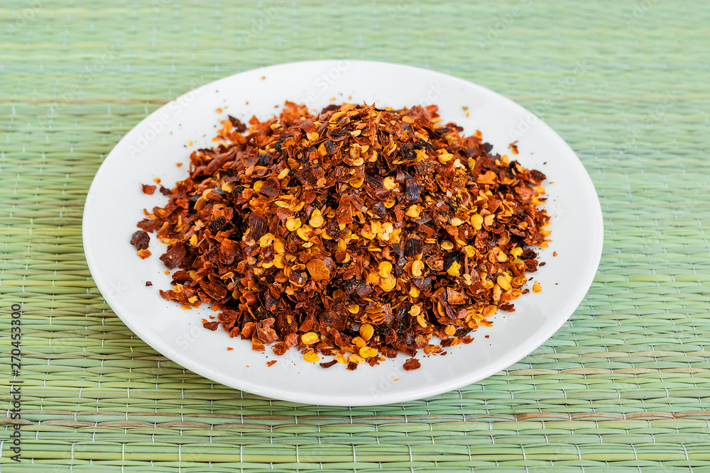 Dried and cruched red hot pepper on a white saucer on a green table mat made of natural plant fibers. Chilli pepper flakes. Natural food spices and seasonings. Dried and cruched red hot pepper on a white saucer on a green table mat made of natural plant fibers. Chilli pepper flakes. Natural food spices and seasonings.