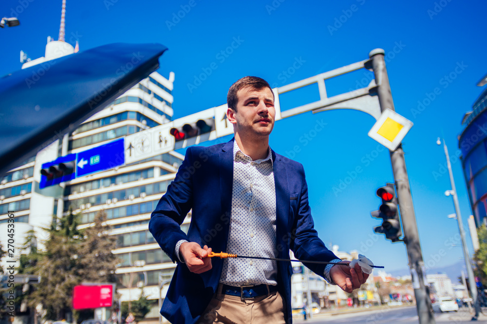 A handsome businessman wearing blue blazer lifting up the hood of his car and checking the oil level on a sunny day parked on a busy city boulevard. A handsome businessman wearing blue blazer lifting up the hood of his car and checking the oil level on a sunny day parked on a busy city boulevard.