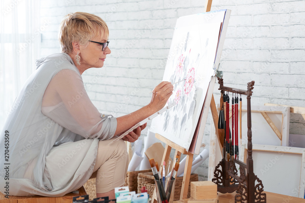 Mature woman sitting in art studio in front of easel and painting a picture with paints - obrazy, fototapety, plakaty 