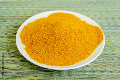 A lot of turmeric or curcuma powder on a white saucer on a green table mat made of natural plant fibers. Natural food spices and seasonings.