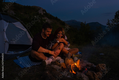 Young couple relaxing by the campfire and drinking coffee in the forest hill in the dusk