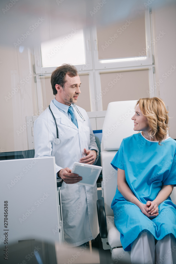 selective focus of handsome doctor holding digital tablet near happy patient sitting in armchair selective focus of handsome doctor holding digital tablet near happy patient sitting in armchair