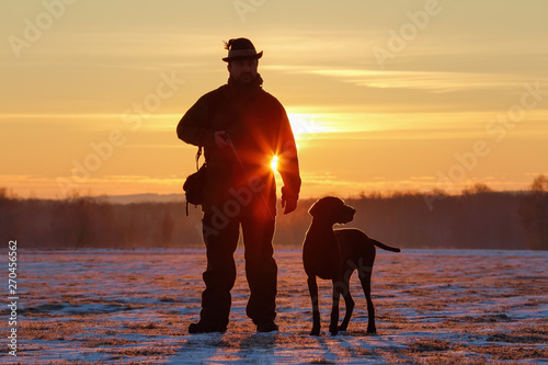 A hunter in the hat with a gun and his German Shorthaired Pointer dog breed friend. Beautiful silhouettes on the background of the dawn. Amazing winter morning. photo
