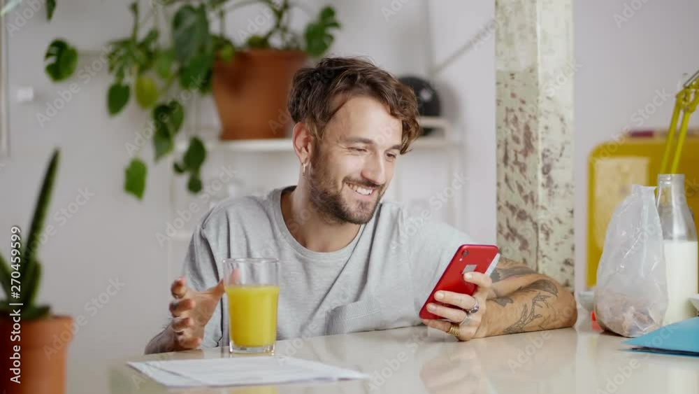 Young cheerful man using red smartphone device drinking fresh orange juice at kitchen , joyful morning routine of tattooed hands male enjoying being online connected in internet with family and Young cheerful man using red smartphone device drinking fresh orange juice at kitchen , joyful morning routine of tattooed hands male enjoying being online connected in internet with family and