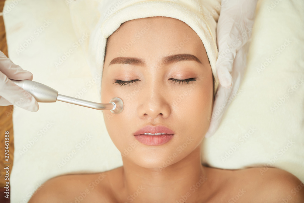 Asian beautiful woman relaxing with eyes closed while cosmetologist in protective gloves doing ultrasound procedure for her face