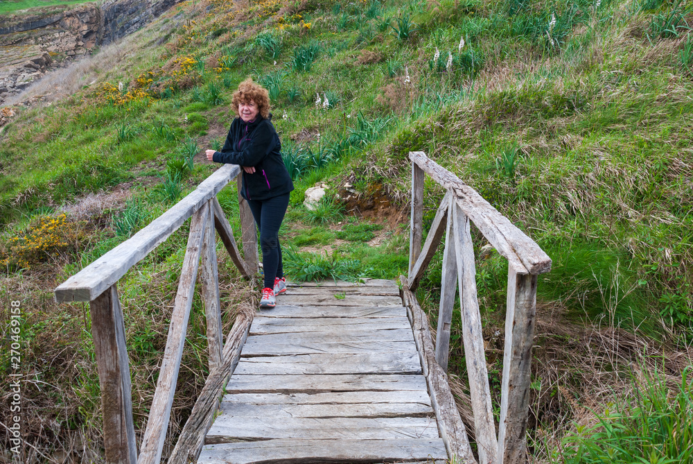 Naklejka premium Mature redhead woman, with sportswear, leaning against a wooden bridge, with meadows in the background, a cloudy spring afternoon