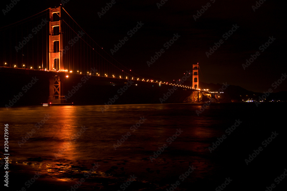 Obraz premium SAN FRANCISCO, CALIFORNIA, UNITED STATES - NOV 25th, 2018: Golden Gate Bridge as seen from Fort Point overlook is glowing in the night with star trails in the sky behind it