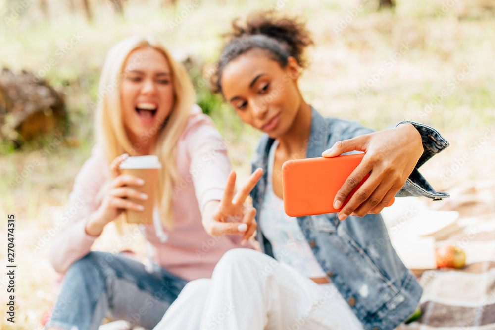 selective focus of two smiling multicultural friends with paper cup of coffee taking selfie and showing peace sign selective focus of two smiling multicultural friends with paper cup of coffee taking selfie and showing peace sign