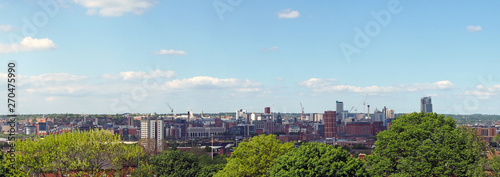 a wide panoramic view showing the whole of leeds city center with towers apartments roads and commercial buildings surrounded by trees against a blue sky