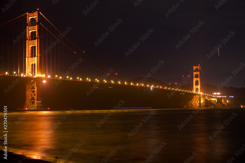 Fototapeta premium SAN FRANCISCO, CALIFORNIA, UNITED STATES - NOV 25th, 2018: Golden Gate Bridge as seen from Fort Point overlook is glowing in the night with star trails in the sky behind it