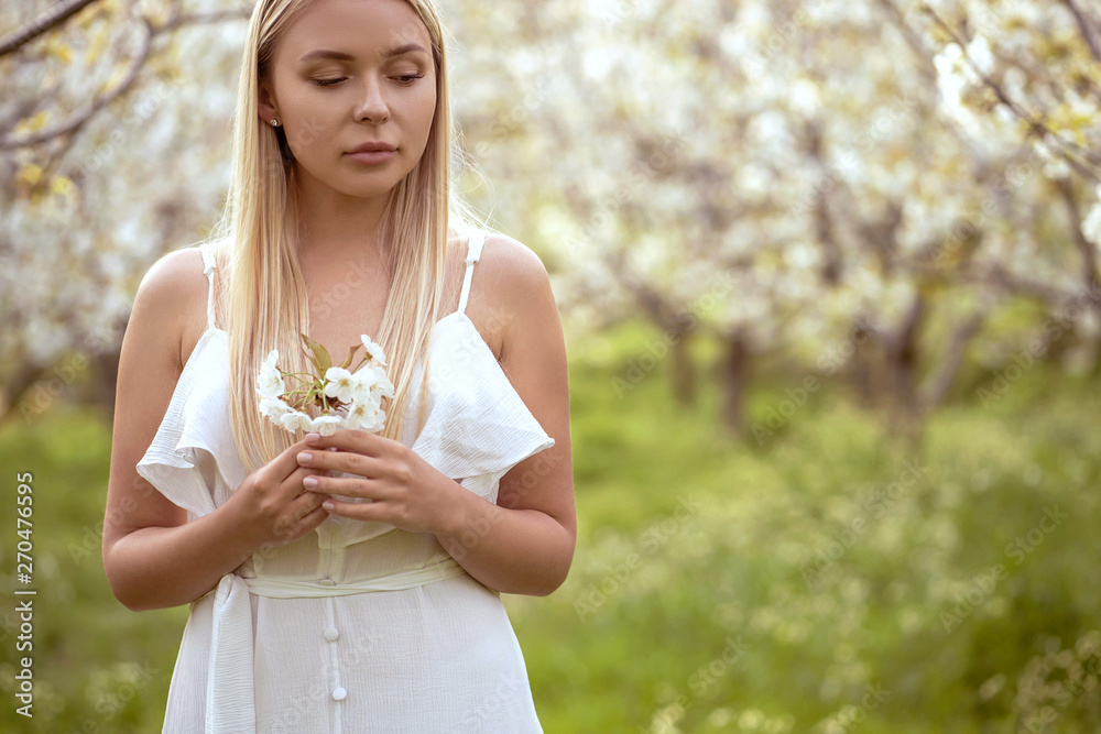 romantic smiling girl in white dress standing at the blossoming garden, holding flower in her hand and looking down; spring season concept. romantic smiling girl in white dress standing at the blossoming garden, holding flower in her hand and looking down; spring season concept.