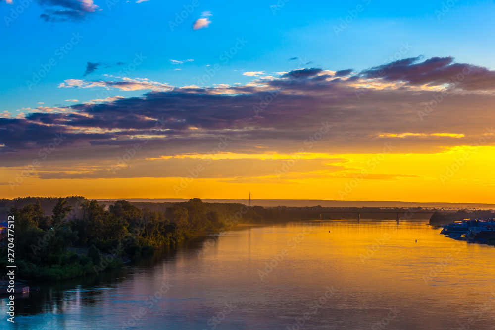 Bright colorful orange sunset over the river, dense dark forest on the shore, railway bridge across the river, reflection of the yellow blue sky in the water. Belaya river, Ufa, Bashkortostan, Russia.