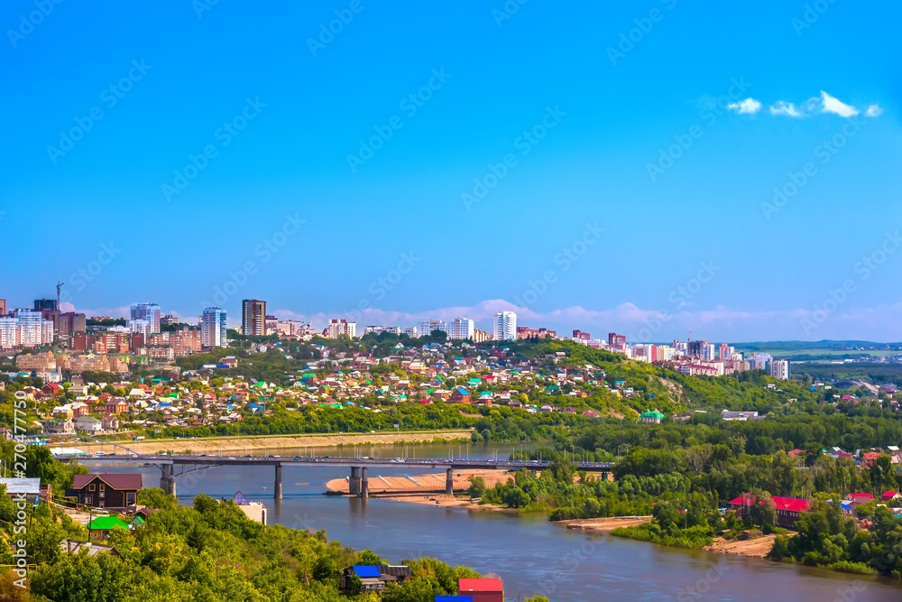 White River in the city center on the background of private houses with colorful roofs on a slope with green trees and modern high-rise buildings. Belsky Bridge, Ufa, Bashkortostan, Russia.