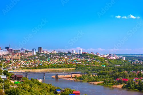 White River in the city center on the background of private houses with colorful roofs on a slope with green trees and modern high-rise buildings. Belsky Bridge, Ufa, Bashkortostan, Russia.