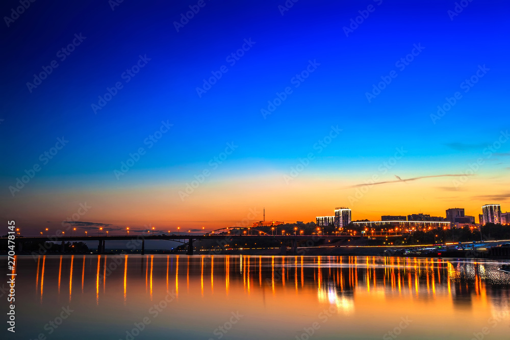 Belsky bridge over the river after sunset at night in the bright orange lights of the city with reflection in the water and a clean colorful blue sky at dusk. White river, Ufa, Bashkortostan, Russia.