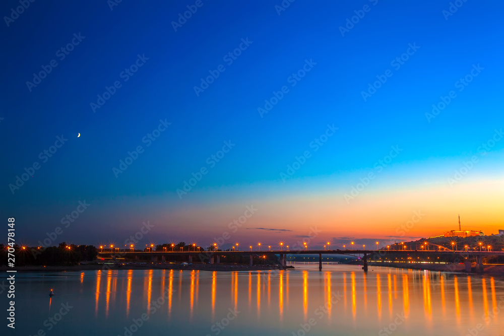Obraz premium Belsky bridge over the river after sunset at night in the bright orange lights of the city with reflection in the water and a clean colorful blue sky at dusk. White river, Ufa, Bashkortostan, Russia.