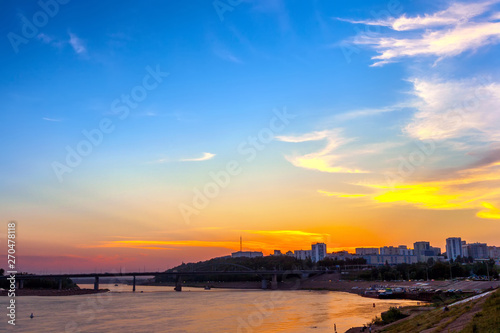 Belsky Bridge over the river at sunset under beautiful clouds of white color with the light of the yellow orange setting sun over the modern city buildings. White river, Ufa, Bashkortostan, Russia.