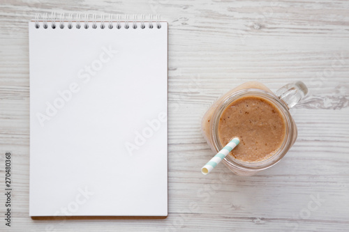 Banana apple smoothie in a glass jar, blank notepad over white wooden background, top view. Flat lay, from above, overhead.