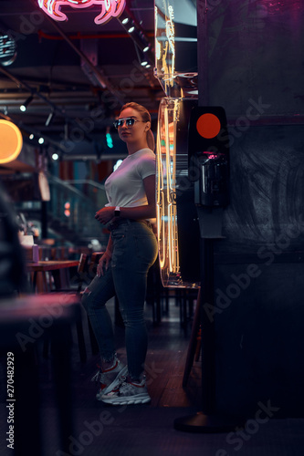 Attractive young woman in sunglasses at neon bar is posing for photographer.
