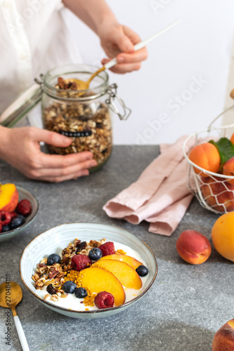 Female hands serving granola from jar, with greek yogurt fresh peach, blueberry, raspberry on the table - healthy breakfast