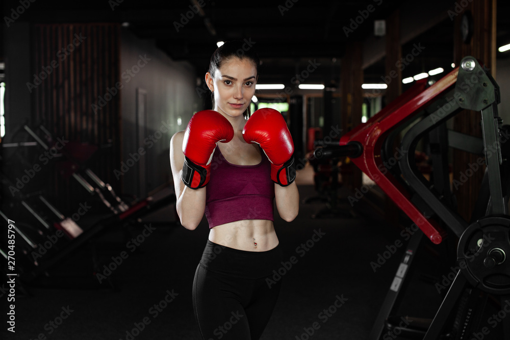 Young pretty woman in red boxing gloves on a background of the gym. Female boxer. Close up portrait of fighter girl. Winner.