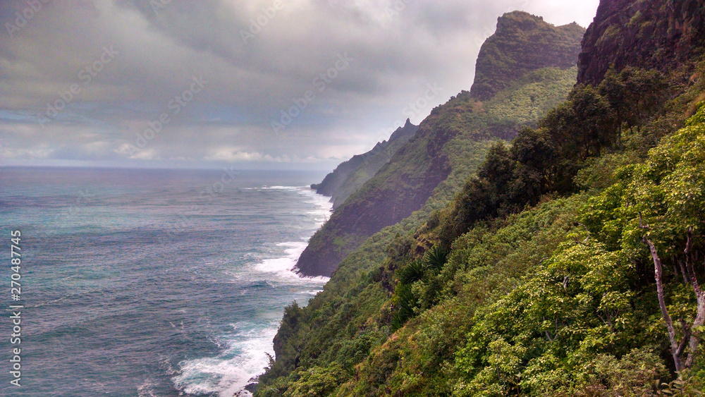 A view of the remote coastline along the Na Pali Coast in Hawaii on the island of Kauai with blue ocean and cloudy sky and lush jungle