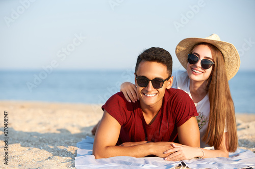 Happy interracial couple newlyweds lie in the sun on tropical travel location. Boyfriend and girlfriend relax at spa resort sunbathing near blue lagoon. Woman with teeth braces wearing straw hat.