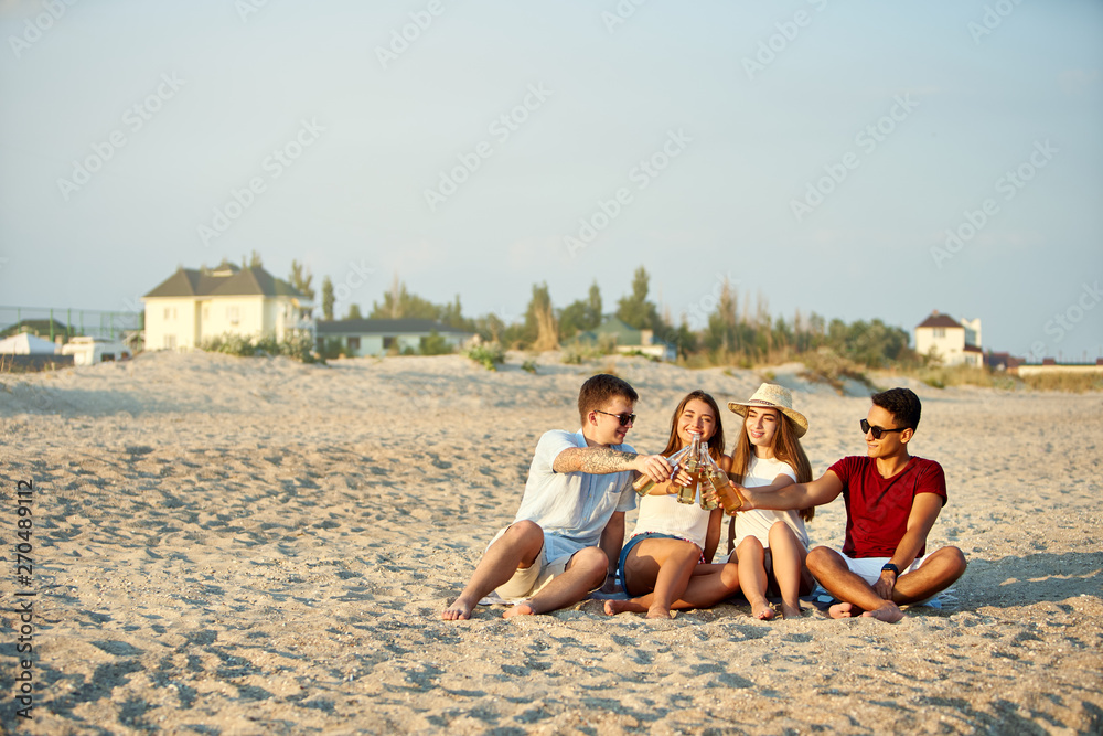Group of friends having fun enjoying a beverage and relaxing on the beach at sunset in slow motion. Young men and women drink beer sitting on a sand in the warm summer evening. Group of friends having fun enjoying a beverage and relaxing on the beach at sunset in slow motion. Young men and women drink beer sitting on a sand in the warm summer evening.