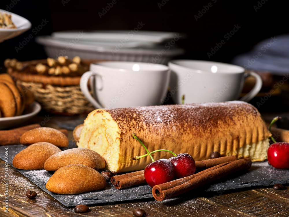 Oatmeal cookies and rolled cake on tier cake stand with cherry and nut on kitchen on wooden table in village style. Two cups of tea on wooden table. Delight pleasant taste. Limitations for diabetics. Oatmeal cookies and rolled cake on tier cake stand with cherry and nut on kitchen on wooden table in village style. Two cups of tea on wooden table. Delight pleasant taste. Limitations for diabetics.