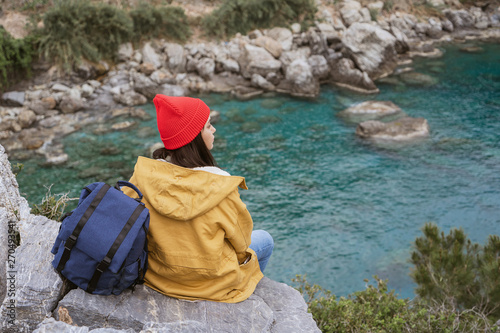 Happy girl with backpack in nature enjoying and sitting on the stone in front of lake, relax time on holiday concept travel, place for text, tone of hipster style.