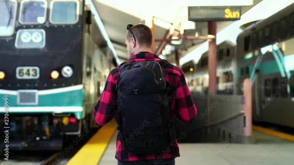 Slow motion: Beautiful young male backpacker walking on platform at Los Angeles train station. Concept of summer adventure, city destination travel, millennial on road trip. 