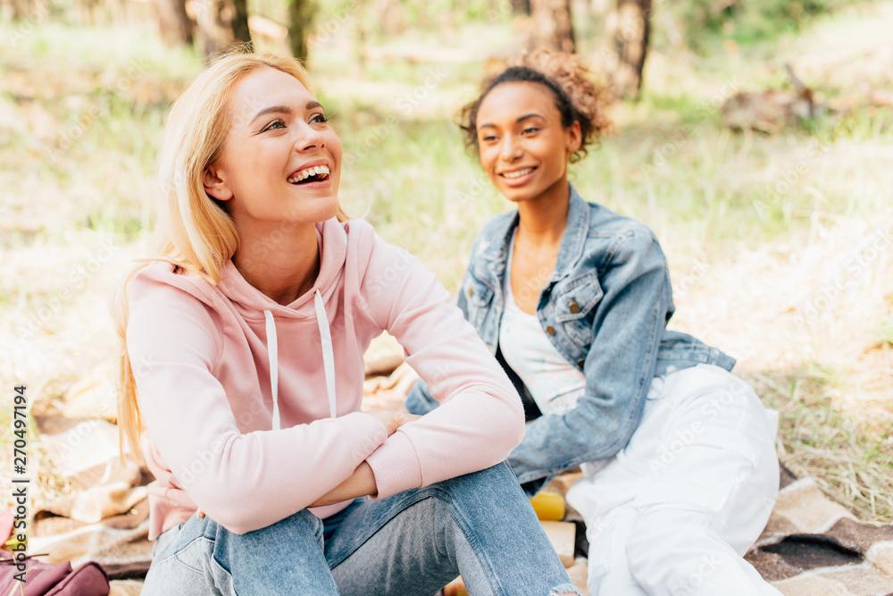 Fototapeta premium two laughing multiethnic friends sitting on plaid blanket in forest