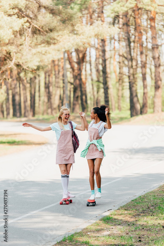 full length view of two smiling multicultural friends holding hands while skateboarding on penny boards on road