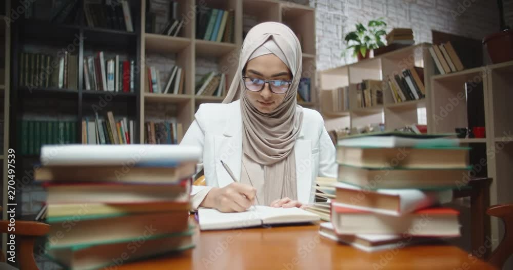 Muslim student is doing research at desk full of books in library. Girl wearing hijab is preparing for exam or making a project, then looking at camera 4k Muslim student is doing research at desk full of books in library. Girl wearing hijab is preparing for exam or making a project, then looking at camera 4k