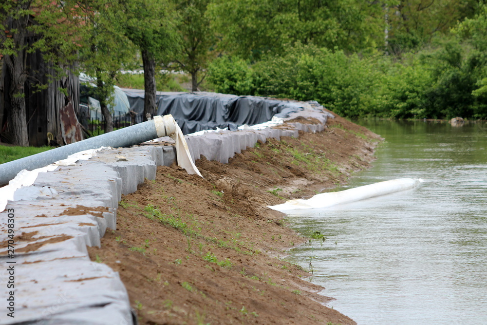 Fototapeta premium Strong metal pipe with connected large diameter fire hose used to pump flood water over wall made of sandbox barriers on rainy spring day