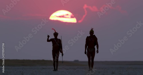 Two Bushman walking aross the Makgadikgadi Pans with the sun setting in the red sky behind them, Botswana photo