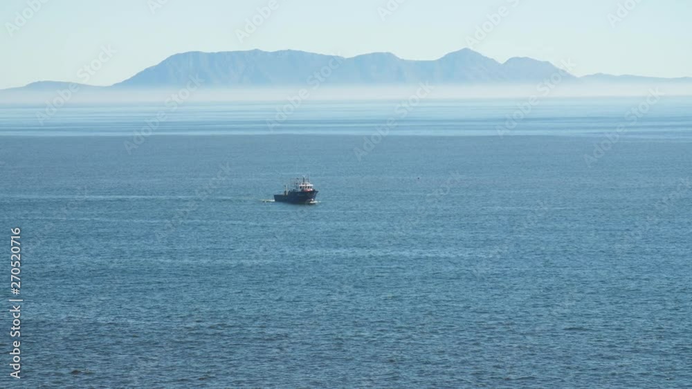 Fishing trawler in distance off coast of South Africa casting fishing nets as it turns in the water with heatwaves visible and misty blue mountains in background on sunny day Fishing trawler in distance off coast of South Africa casting fishing nets as it turns in the water with heatwaves visible and misty blue mountains in background on sunny day