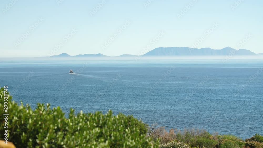 Fishing trawler sailing away in blue waters off the coast of South Africa on clear sunny day with heatwaves and green shrubbery in foreground and misty mountains in background