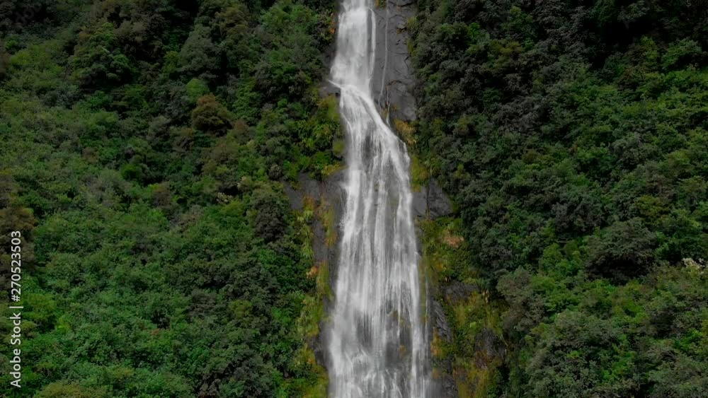 Stunning aerial drone pan shot showing high cliffs covered with dense trees with a waterfall coming down in New Zealand Stunning aerial drone pan shot showing high cliffs covered with dense trees with a waterfall coming down in New Zealand