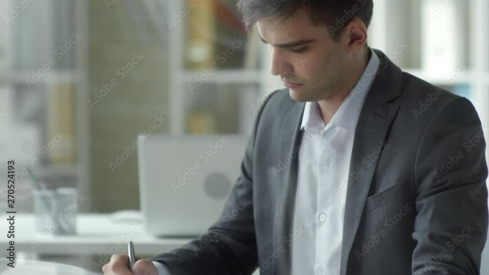Tilt up shot of young caucasian businessman sitting at office desk, using digital tablet and writing down in notepad Tilt up shot of young caucasian businessman sitting at office desk, using digital tablet and writing down in notepad